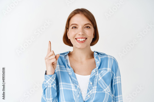 Portrait of happy woman smiling, pointing finger up, showing direction. Young girl announcing special deal, advertising on white background