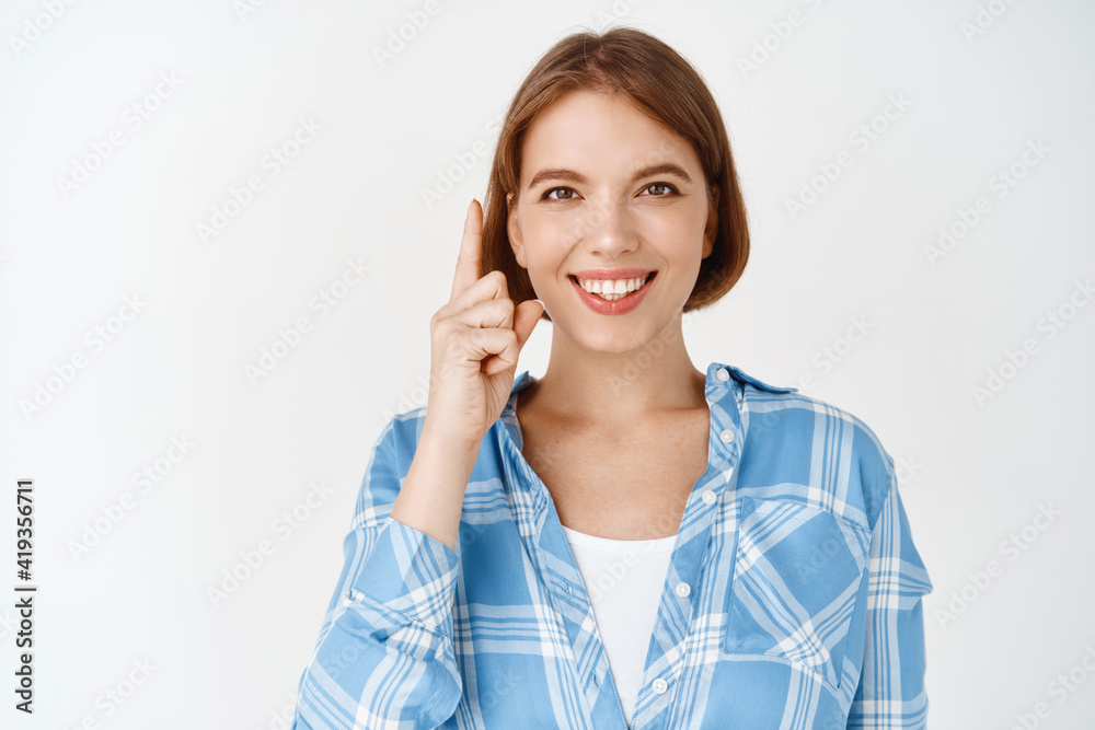 Beautiful young girl with short hair and confident smile, pointing finger up at promo deal, showing advertisement on top and looking happy, white background Beautiful young girl with short hair and confident smile, pointing finger up at promo deal, showing advertisement on top and looking happy, white background