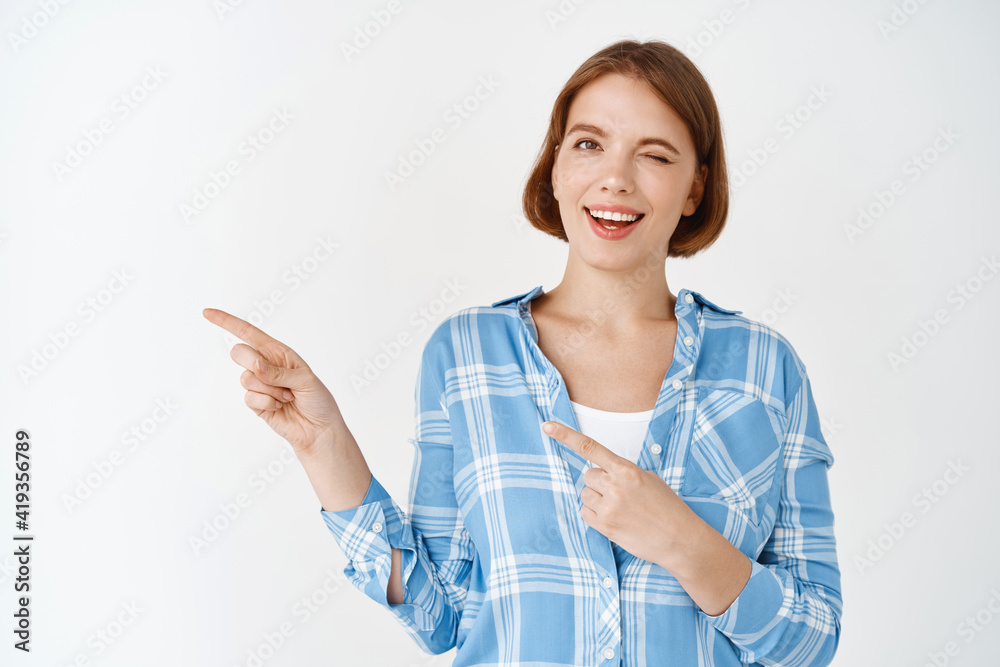 Look here. Smiling natural young woman winking, pointing fingers left at copy space, showing promo special deal, standing on white background Look here. Smiling natural young woman winking, pointing fingers left at copy space, showing promo special deal, standing on white background