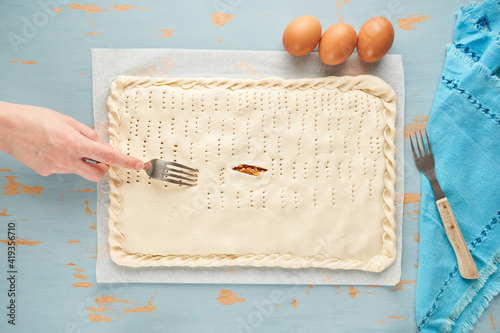 One person pricks the dough with a fork over the tuna, tomato, onion and bell pepper filling to prepare an empanada on a blue wooden table. Tuna pie. Typical Galician dish from Galicia and Spain