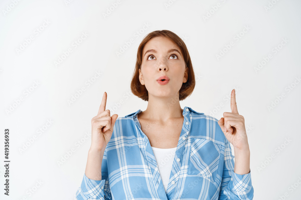 Wow look here. Excited young woman looking and pointing up, pucker lips amazed, checking out promo, standing on white background Wow look here. Excited young woman looking and pointing up, pucker lips amazed, checking out promo, standing on white background
