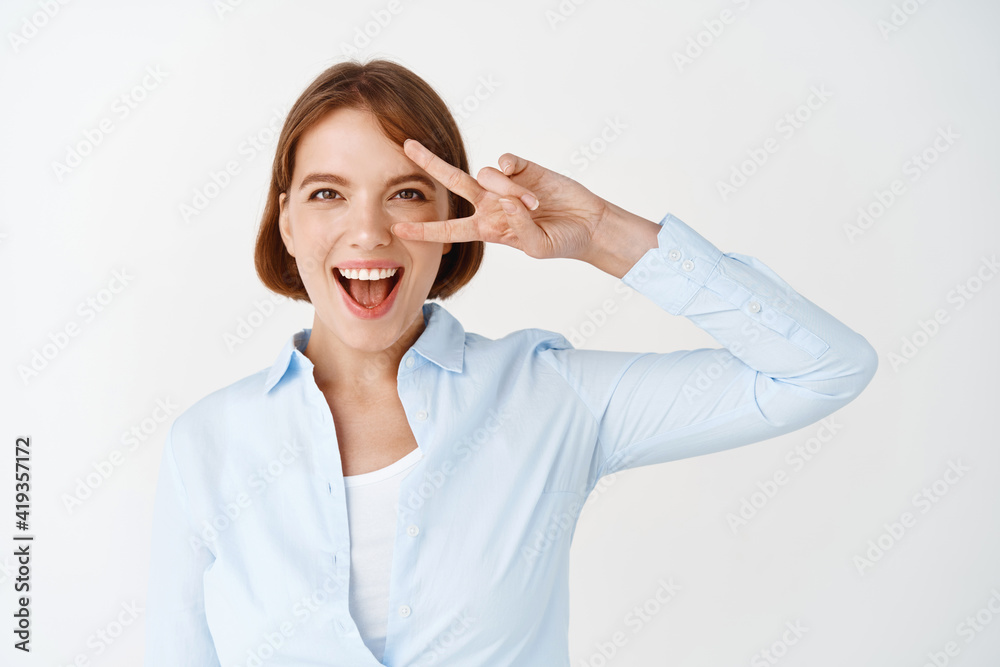 Portrait of excited female office worker, showing v-sign over eye and smiling happy, standing in blouse, feeling lucky and motivated, white background Portrait of excited female office worker, showing v-sign over eye and smiling happy, standing in blouse, feeling lucky and motivated, white background