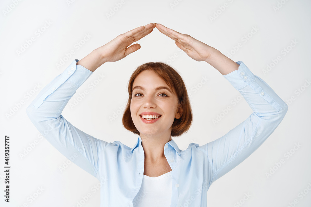 Insurance and real estate concept. Smiling woman in blouse making hand roof about head, showing home gesture, standing on white background Insurance and real estate concept. Smiling woman in blouse making hand roof about head, showing home gesture, standing on white background