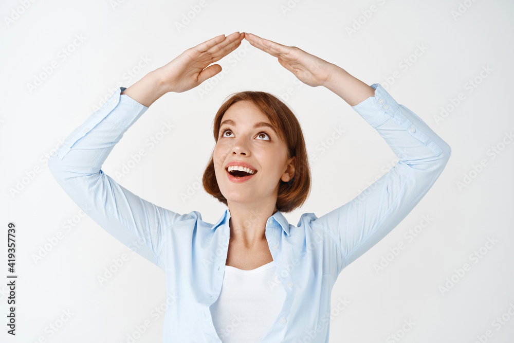 Insurance and real estate concept. Excited young girl looking at hands making roof, showing rooftop of home and smiling, standing against white background Insurance and real estate concept. Excited young girl looking at hands making roof, showing rooftop of home and smiling, standing against white background