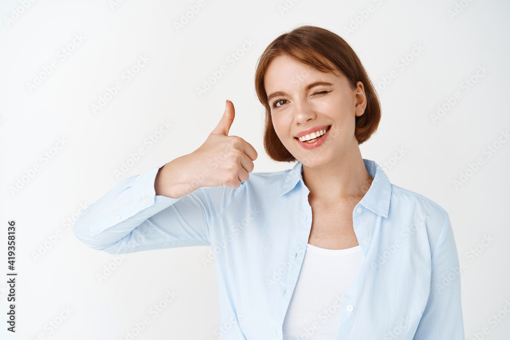 Well done. Smiling successful businesswoman winking, say yes and showing thumb up in approval, like something good, recommending product, standing on white background Well done. Smiling successful businesswoman winking, say yes and showing thumb up in approval, like something good, recommending product, standing on white background