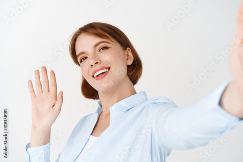 Portrait of woman waving hand to say hi on video chat, holding smartphone in stretched out hand, greeting friend, standing against white background