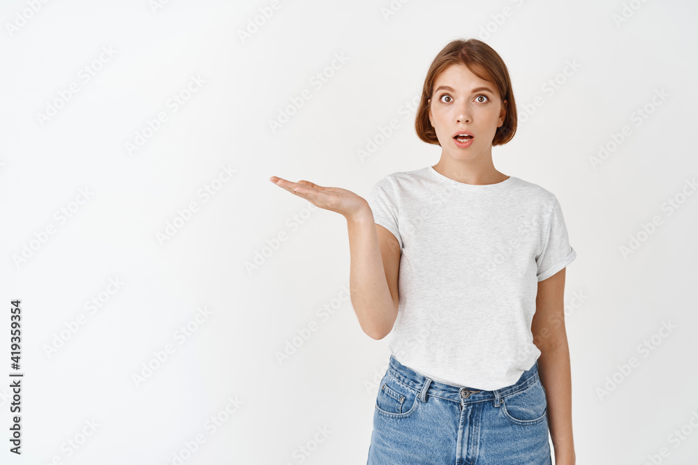 Excited female student holding empty space on palm, holding something in hand and look amazed at camera, standing on white background Excited female student holding empty space on palm, holding something in hand and look amazed at camera, standing on white background