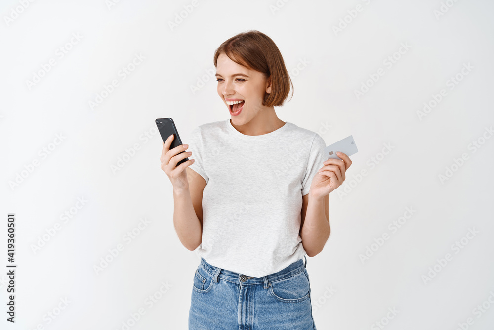 Happy smiling woman holding smartphone and plastic credit card, looking at mobile phone screen excited, making money online, standing against white background Happy smiling woman holding smartphone and plastic credit card, looking at mobile phone screen excited, making money online, standing against white background