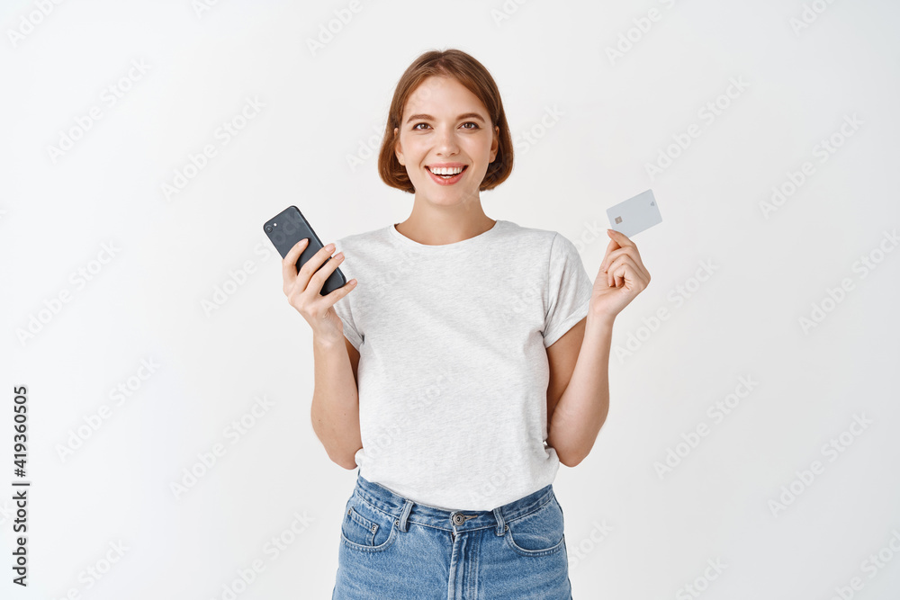 Portrait of smiling natural girl showing mobile phone and plastic credit card, paying online, standing against white background Portrait of smiling natural girl showing mobile phone and plastic credit card, paying online, standing against white background
