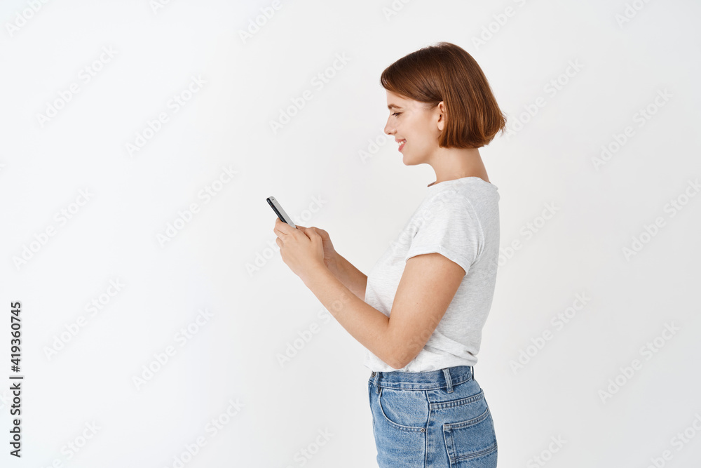 Profile of young woman chatting on mobile phone. Happy girl with short hair texting message with smartphone, standing against white background Profile of young woman chatting on mobile phone. Happy girl with short hair texting message with smartphone, standing against white background