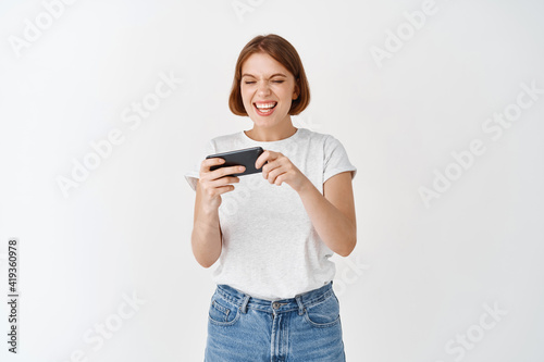 Excited woman playing video game on smartphone, relaxing with mobile phone, standing in t-shirt and jeans against white background