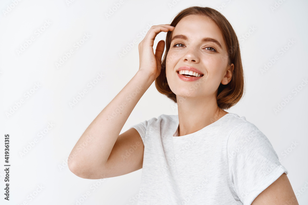 Portrait of happy young woman with natural face light make-up, touching haircut and smiling at camera, standing in t-shirt against white background Portrait of happy young woman with natural face light make-up, touching haircut and smiling at camera, standing in t-shirt against white background