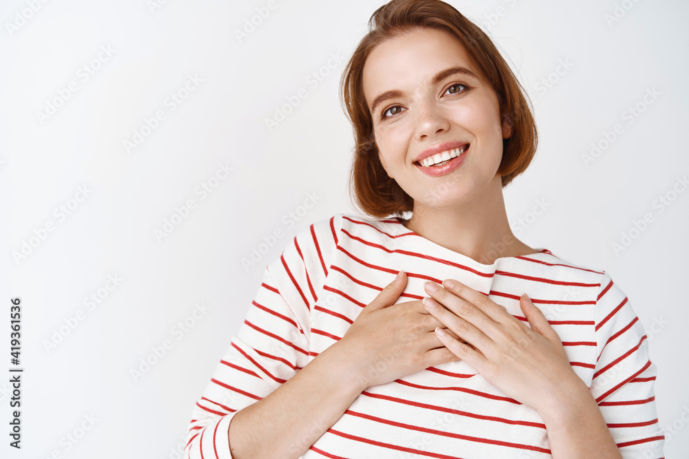 Lovely young woman with short hair, holding hands on heart and smiling thankful, being touched with nice gesture, standing on white background