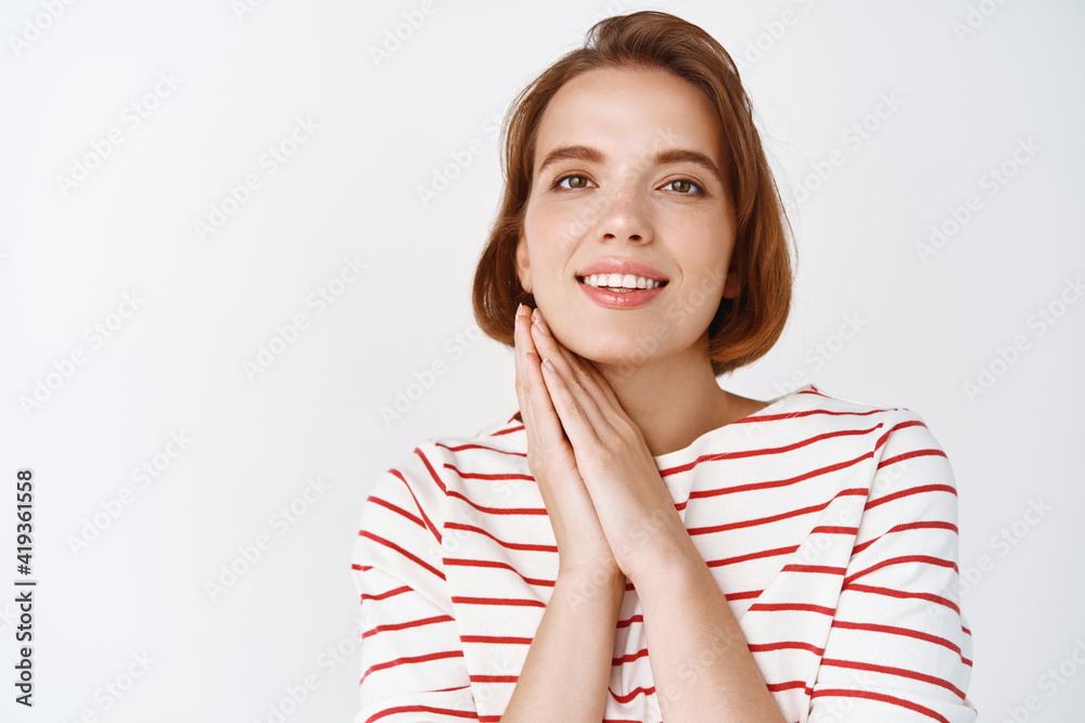 Beauty face. Tender young woman with short hair, holding hands near cheeks, natural skin, smiling at camera, standing in casual clothes against white background