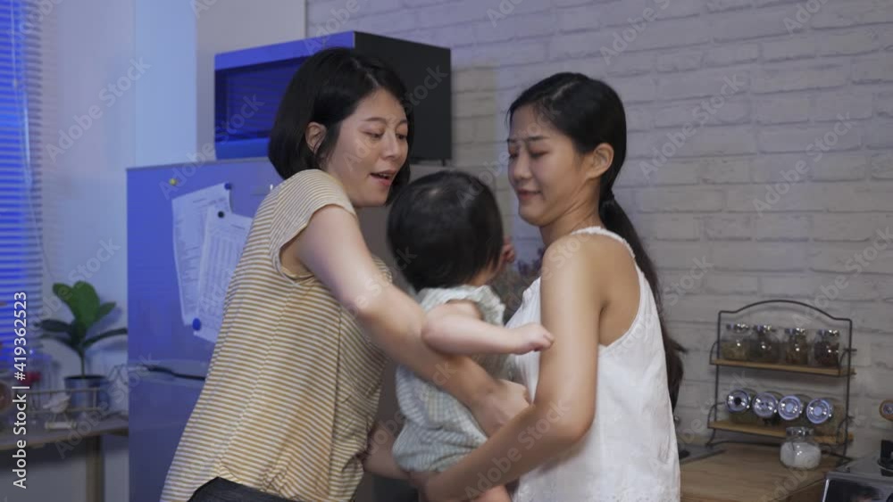 two happy mothers of an lgbt family are taking turns holding their adopted daughter and moving their gaze with her while pointing up and ahead at home two happy mothers of an lgbt family are taking turns holding their adopted daughter and moving their gaze with her while pointing up and ahead at home