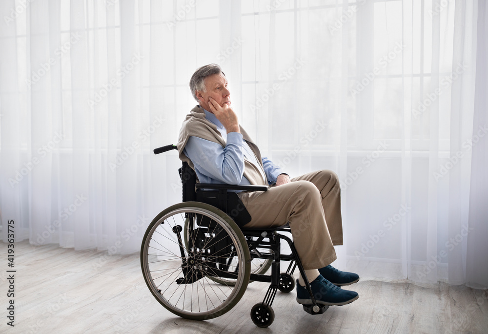 Full length portrait of handicapped senior man in wheelchair feeling desperate and lonely, looking out window indoors Full length portrait of handicapped senior man in wheelchair feeling desperate and lonely, looking out window indoors