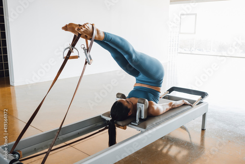 Rear view of a fit woman lying down on a bed of a Pilates Reformer Machine Bed, with legs up, working out and training in in blue workout clothing, at a gym studio. Day light coming from windows.