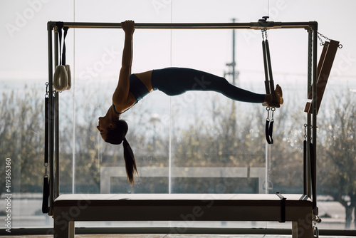 Fit female coach hanging hands and feet from the Pilates machine, woman working out on the Trapeze Table in a big Fitness Gym,  big windows and street views. Pilates and healthy lifestyle concept