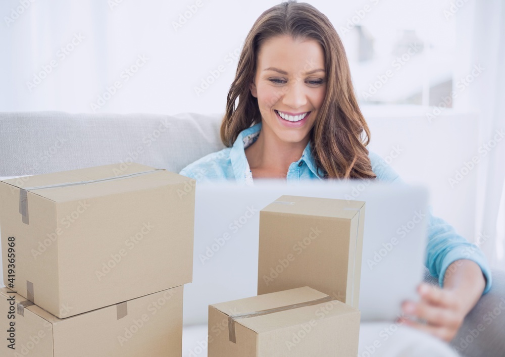 Composition of cardboard boxes with smiling woman using laptop at home Composition of cardboard boxes with smiling woman using laptop at home