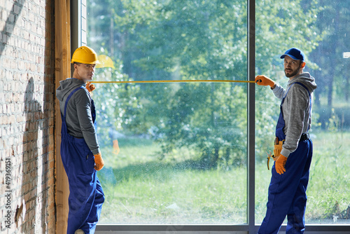 фотографія Two positive young male builders wearing blue overalls looking at camera, using