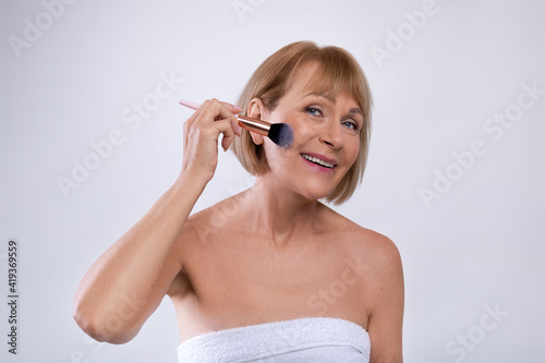 Organic makeup for mature skin. Sensual senior lady putting on blusher or powder on her face over light background