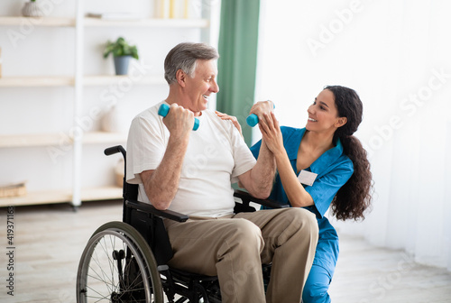 Doctor helping disabled elderly man in wheelchair to work out with dumbbells, to recover from injury at health centre