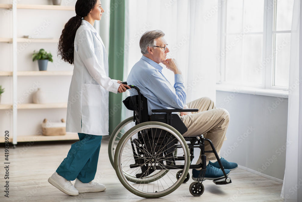 Side view of young doctor helping elderly disabled man in wheelchair, indoors Side view of young doctor helping elderly disabled man in wheelchair, indoors