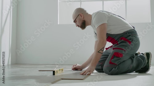 Bald handyman in workwear uses level to check parquet floor