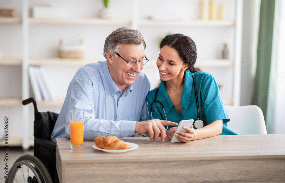 Disabled senior man and young nurse using smartphone together, browsing web or watching movie indoors Disabled senior man and young nurse using smartphone together, browsing web or watching movie indoors
