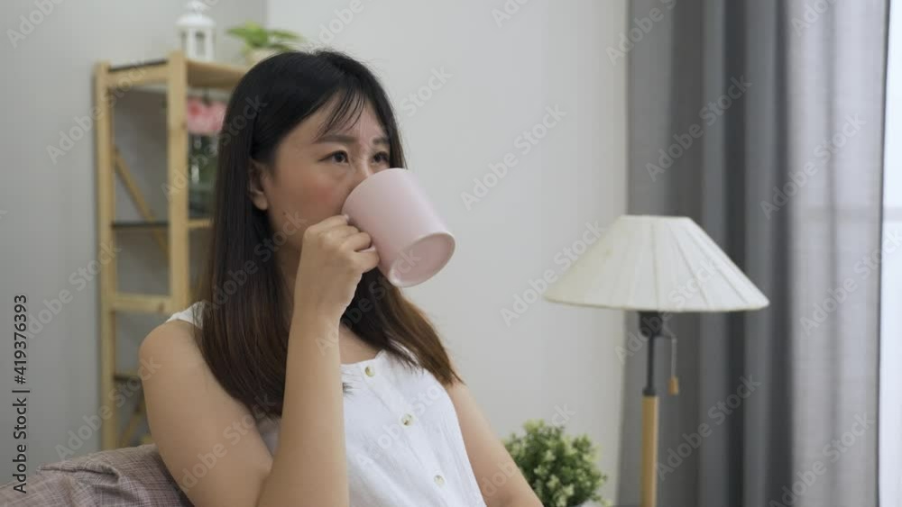 lonely japanese girl seated on the couch in low spirit is drinking tea and looking into the distance with a thoughtful look at a bright home interior. lonely japanese girl seated on the couch in low spirit is drinking tea and looking into the distance with a thoughtful look at a bright home interior.
