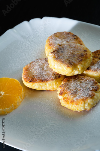 Delicious cottage cheese pancakes with powdered sugar, with an orange on a white plate close-up. Vertical photo