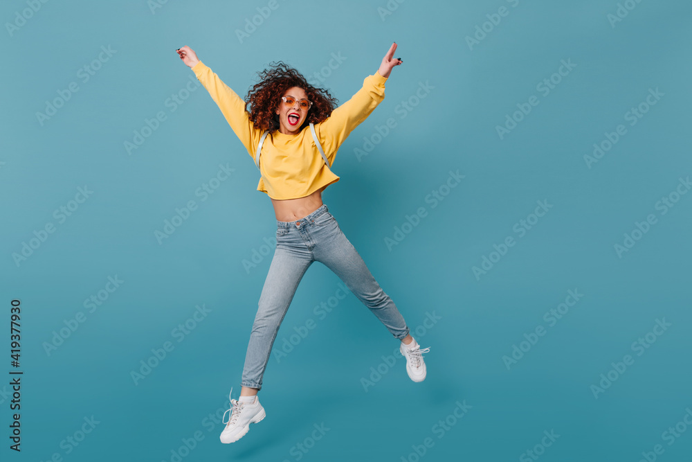 Active girl shows tongue. Woman in yellow sweater and jeans happily jumping on isolated blue background