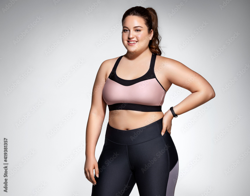 Woman with curvy figure in fashionable sportswear on grey background. Sports motivation and healthy lifestyle