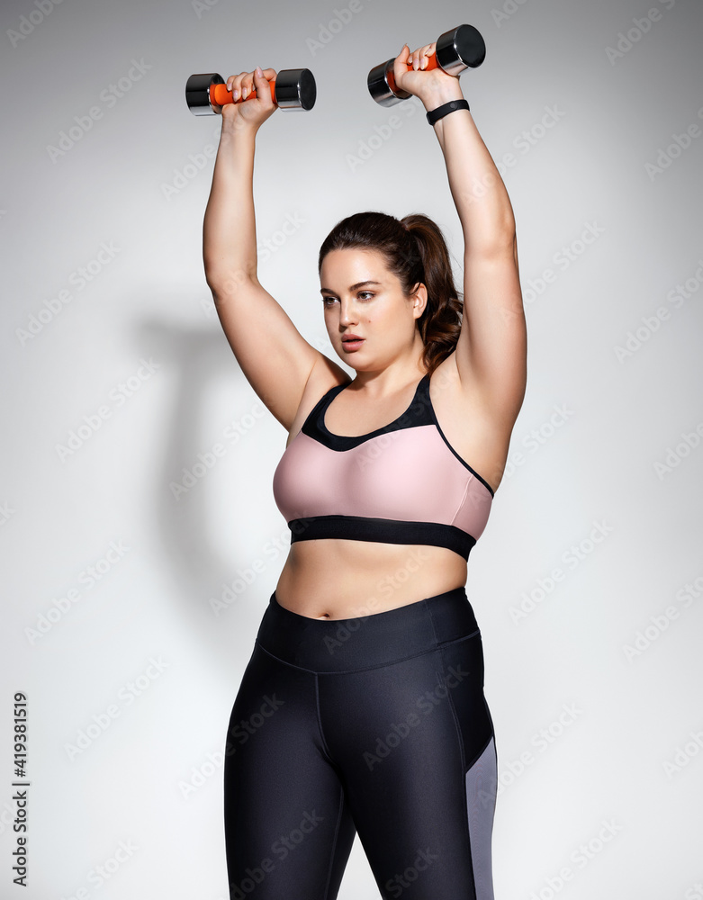 Sporty woman doing exercises with dumbbells. Photo of model with curvy figure in fashionable sportswear on grey background. Sports motivation and healthy lifestyle Sporty woman doing exercises with dumbbells. Photo of model with curvy figure in fashionable sportswear on grey background. Sports motivation and healthy lifestyle