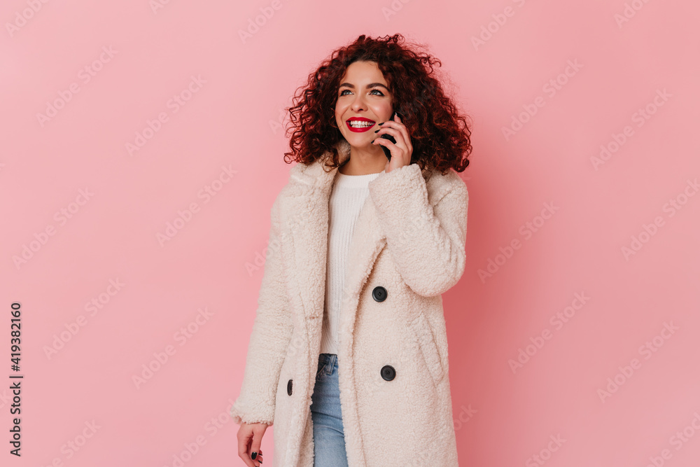 Stylish girl in eco-coat and jeans talking on phone. Woman with red lips and white smile posing over pink background Stylish girl in eco-coat and jeans talking on phone. Woman with red lips and white smile posing over pink background
