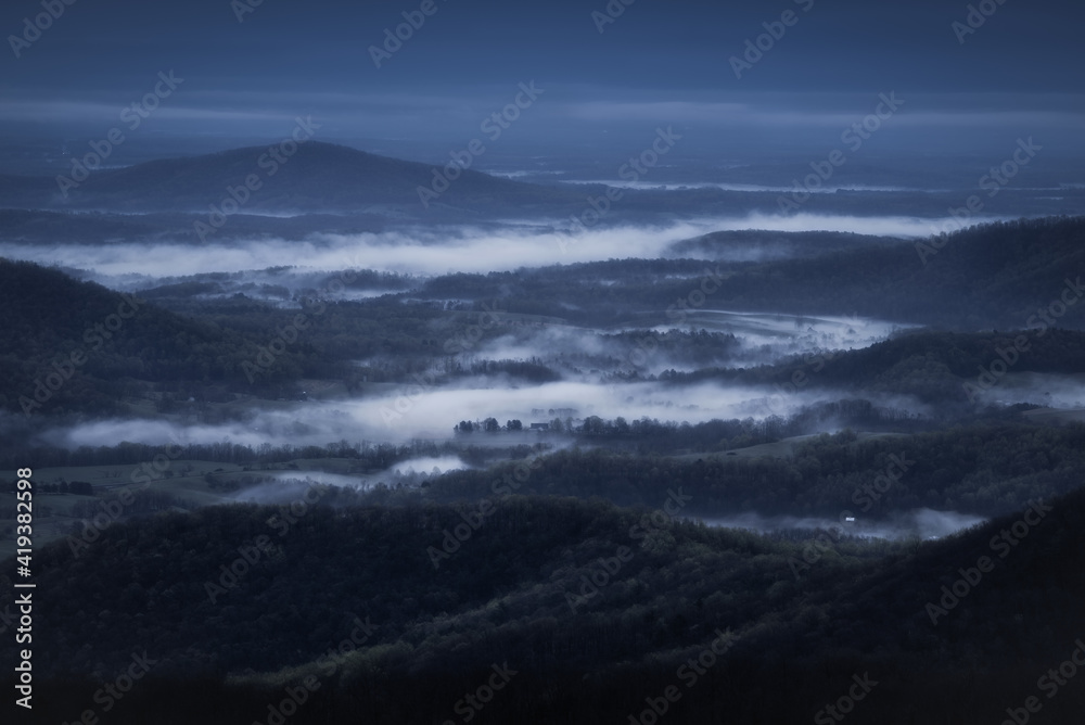 White mist fills the rural countryside of Virginia on a dark and moody, blue morning viewed from Shenandoah National Park.