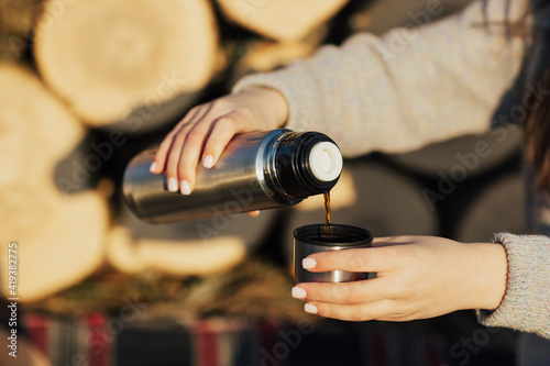 Close up of hands of girl pouring the hot drink from a thermos while sitting near pile of wood logs in forest. Picnic in forest.