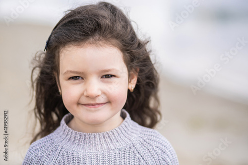 Closeup view portrait of cute calm pretty white smiling happy little girl with long brown hair standing outdoor on sunny spring sandy beach. Sweet child looking in camera happily