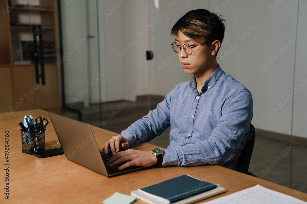 Focused asian guy working with laptop while sitting at table in office Focused asian guy working with laptop while sitting at table in office