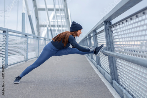 Young happy fitness woman in winter blue sportswear stretch legs after jogging on bridge metal fence during cold day