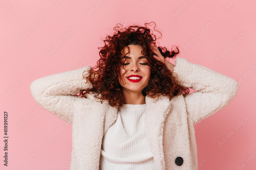 Portrait of charming lady with red lipstick and snow-white smile dressed in bright eco-coat and touching her curly hair on pink background Portrait of charming lady with red lipstick and snow-white smile dressed in bright eco-coat and touching her curly hair on pink background