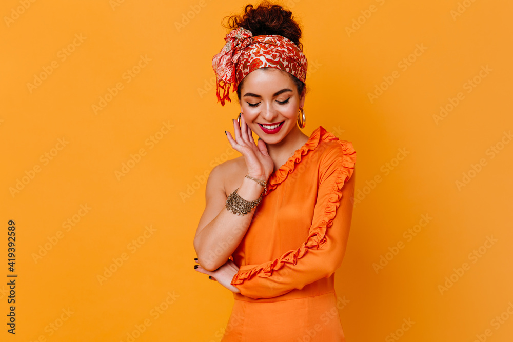 Stylish lady in orange dress and bright bandage on her head with shy smile looks down on orange background Stylish lady in orange dress and bright bandage on her head with shy smile looks down on orange background