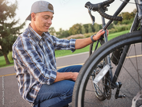 Portrait of joyful young guy in casual wear smiling while repairing his bike, kneeling on the road in public park