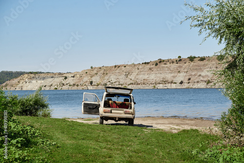 Scenic view over wide river with old car, parked on it's bank. Beautiful natural countryside landscape with blue sunny sky. Summer getaway vacation into isolated places.