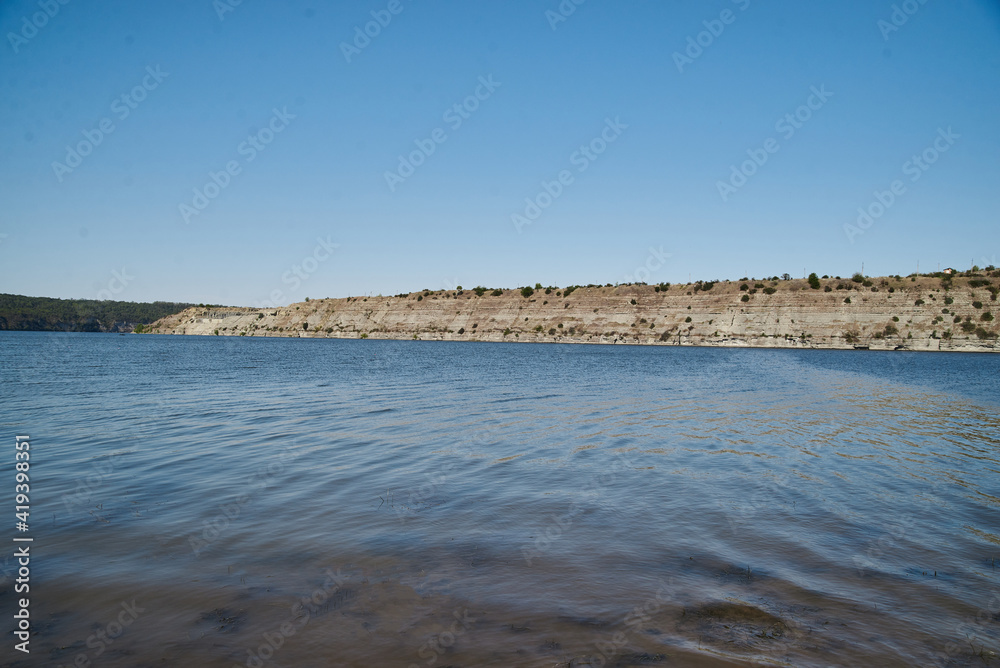 Obraz premium Scenic view over wide river and it's bank. Beautiful natural countryside landscape with blue sunny sky. Summer getaway vacation into isolated places. Calm water - perfect for meditation.