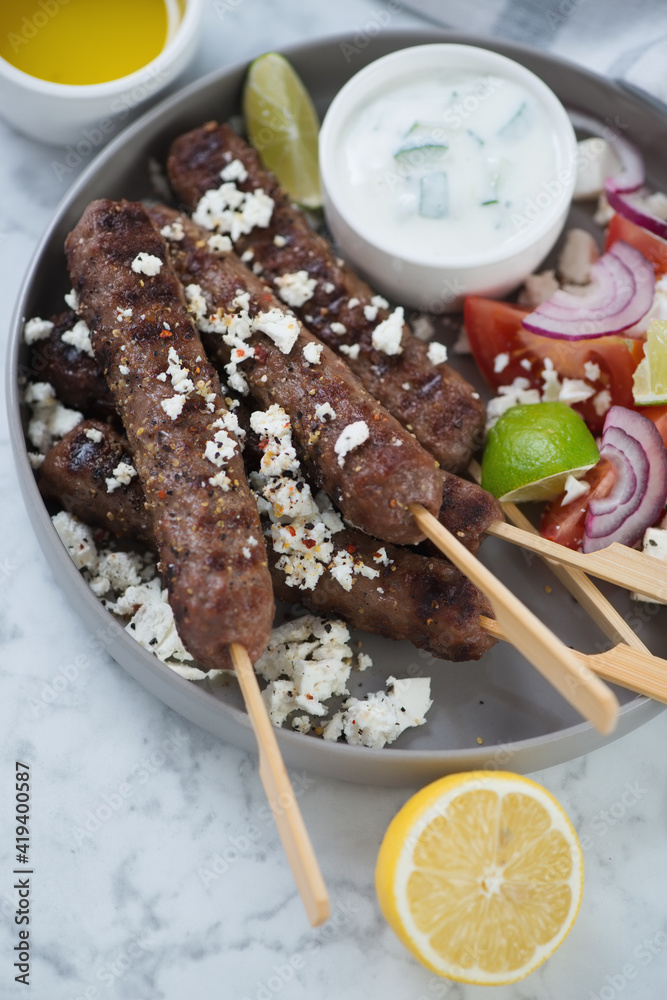 Fototapeta premium Closeup of roasted greek kofta kebabs with served with feta cheese and tzatziki sauce, vertical shot on a grey marble surface