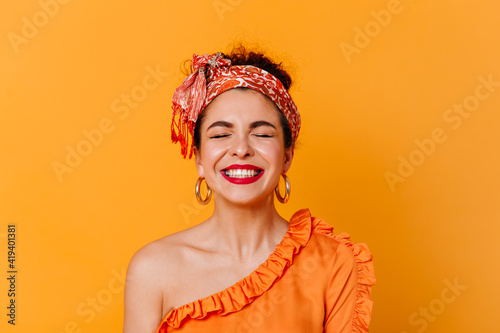 Close-up portrait of positive woman with red lips dressed in blouse with bare shoulder and headband laughing with closed eyes on isolated background