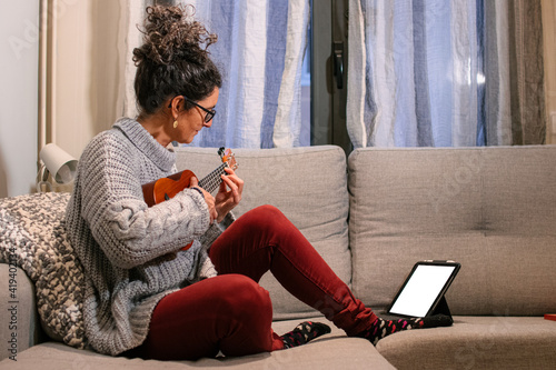 Mujer caucásica de mediana edad sentada en el sofa con ropa casual aprendiendo a tocar el ukelele por internet a traves de una tablet.