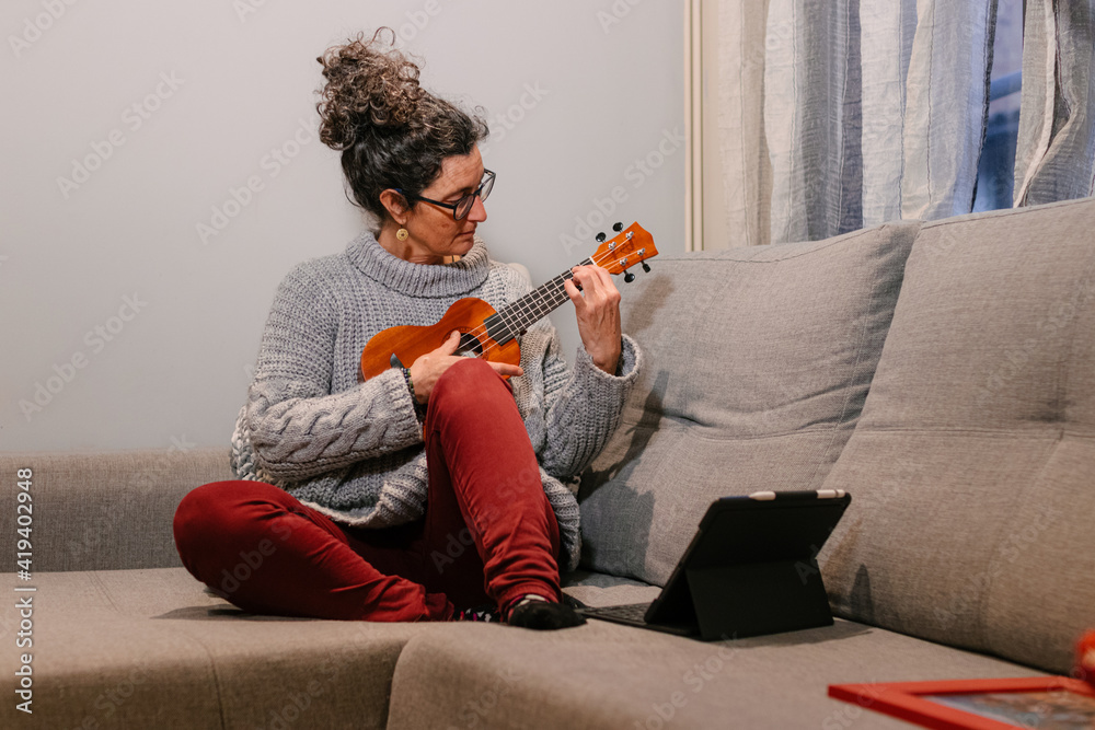 Mujer caucásica de mediana edad sentada en el sofa con ropa casual aprendiendo  a tocar el ukelele por internet a traves de una tablet. - obrazy, fototapety, plakaty 