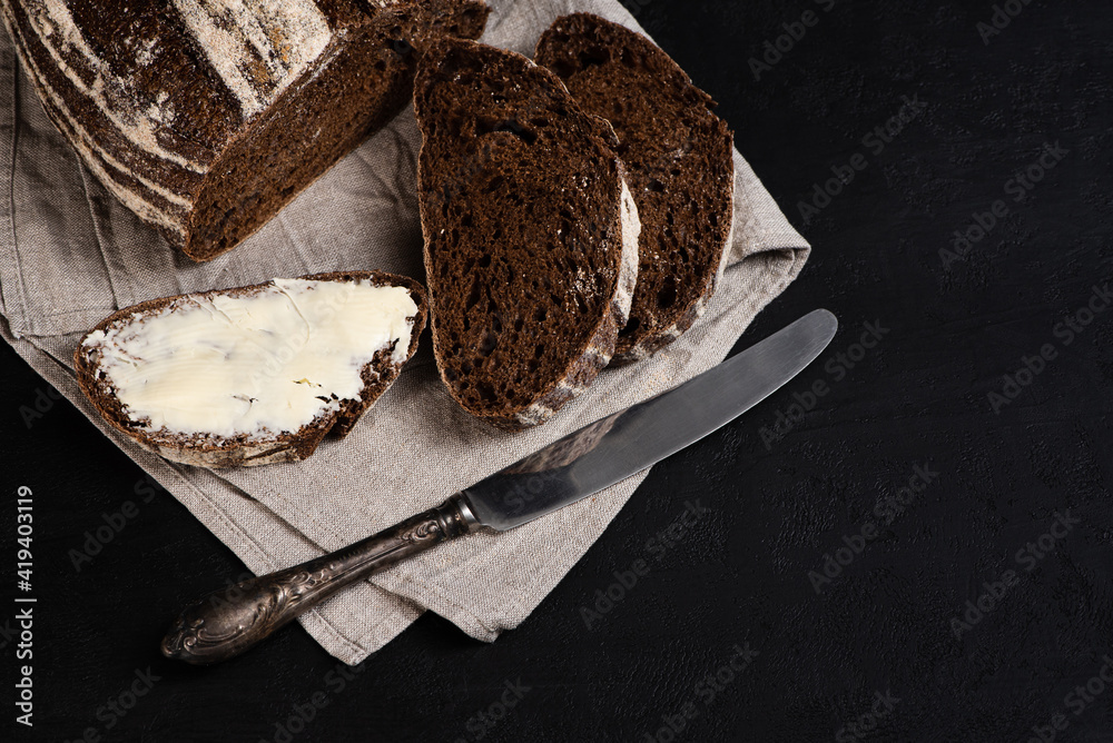 Buttered slice of freshly baked rye bread, table knife on dark background. Artisan sourdough bread. Copy space for recipe. View from above.